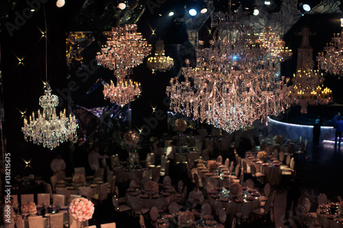 Luxury large crystal chandeliers hang over round dinner tables prepared for the party