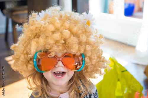 funny portrait of three years old child disguised as sixties, with great curly blond hair wig with daisy flowers on head, with orange and green colorful glasses, looking laughing