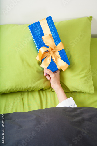 woman hand coming out of blue and green sheets with paper wrapped gift box with ribbon over pillow 