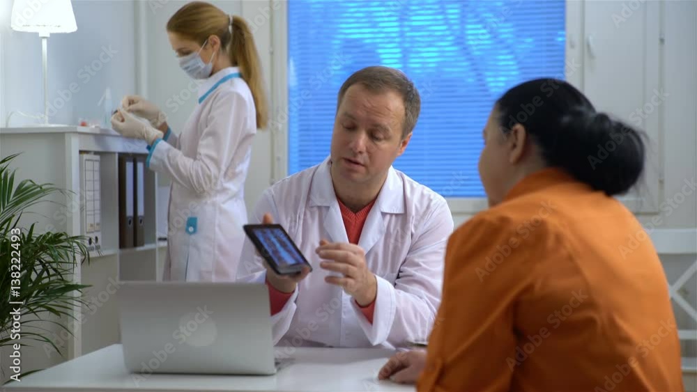 Handsome medical doctor is talking to female and showing her a X-ray picture on tablet while working in his office