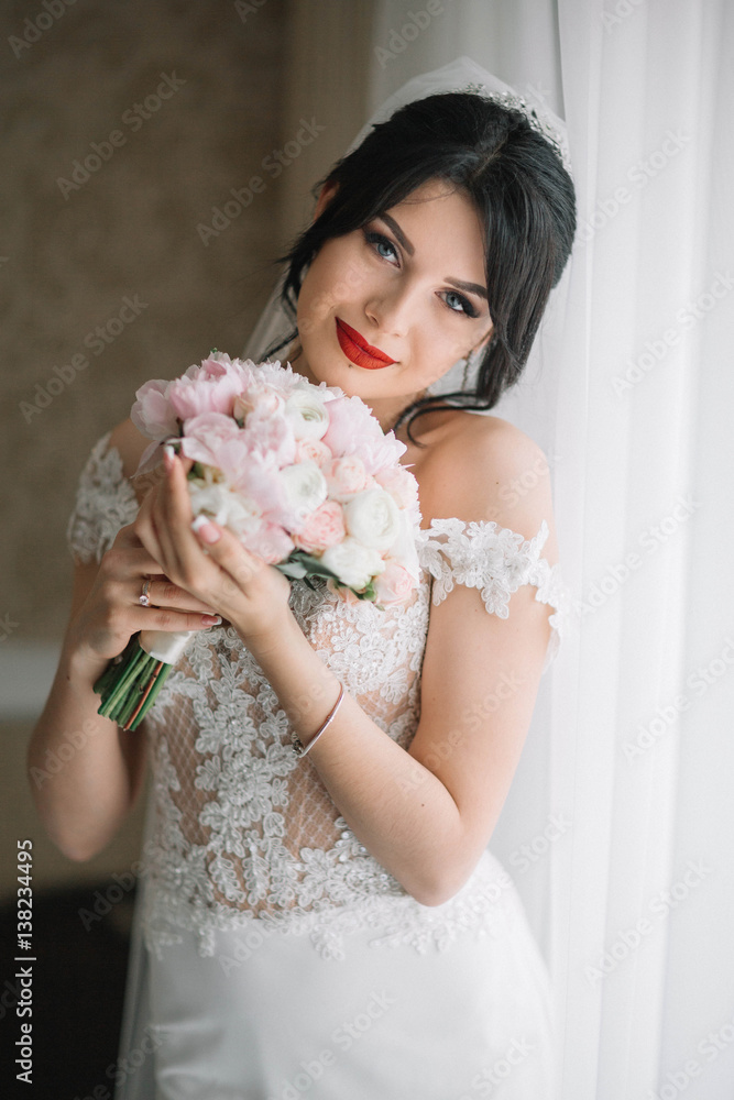 Naklejka premium beautiful and young bride with bouquet of flowers standing near window