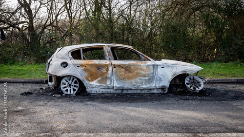 A burnt-out car left abandoned at the road side of a quiet rural location.