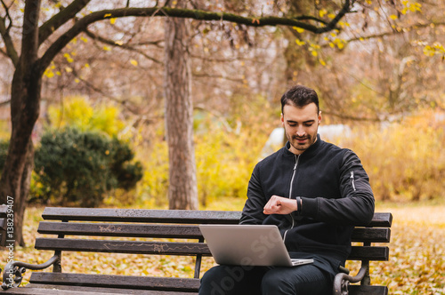 Young man sitting on bench with laptop and looking at watch