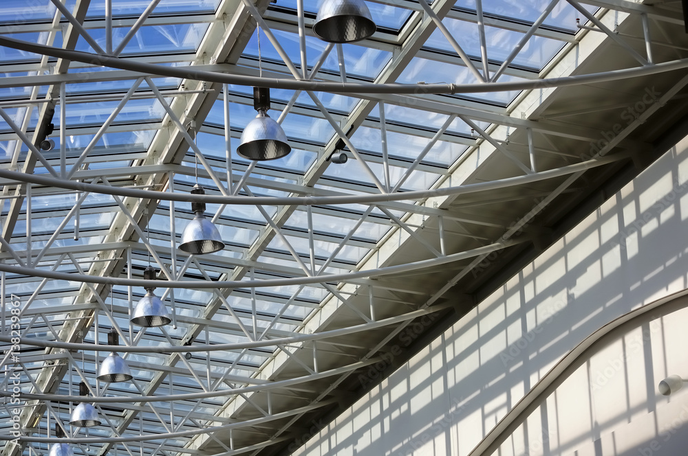 Fragment of modern building with roof made of glass and metal with row of lamps against blue sky background
