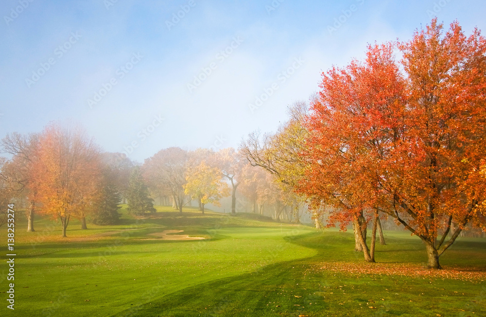 Misty autumn morning. Morning mist and sun light. Fog over golf course during beautiful fall sunrise. Colorful autumn trees on a bright green lawn. Horizontal composition.