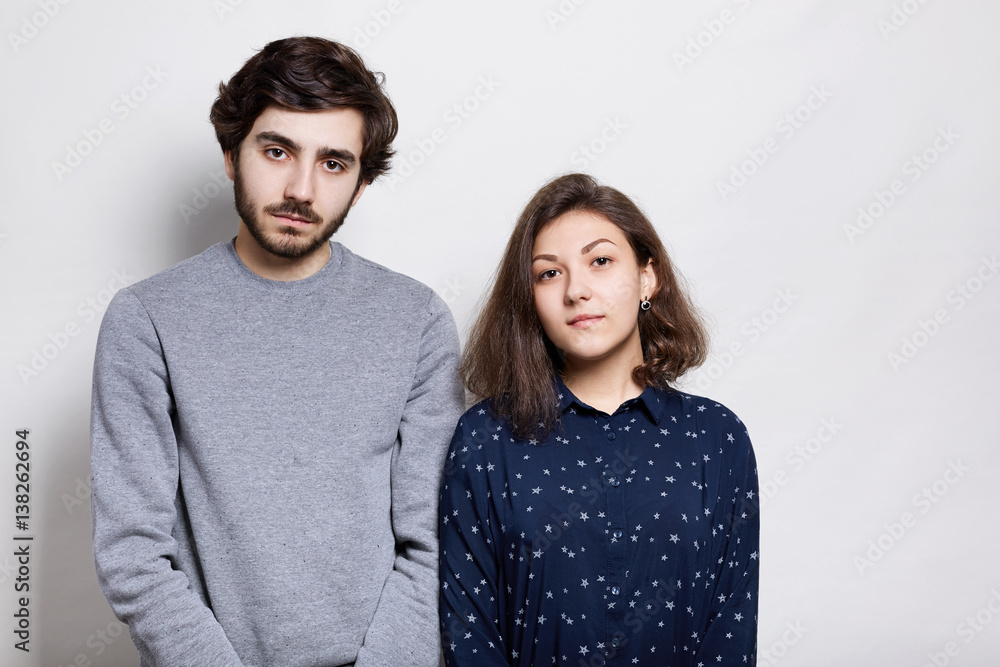 Cozy shot of two beautiful friends standing near each other. Hipster with trendy beard and mustache dressed in gray sweater and pretty girl in dark shirt having mysterious expression Cozy shot of two beautiful friends standing near each other. Hipster with trendy beard and mustache dressed in gray sweater and pretty girl in dark shirt having mysterious expression