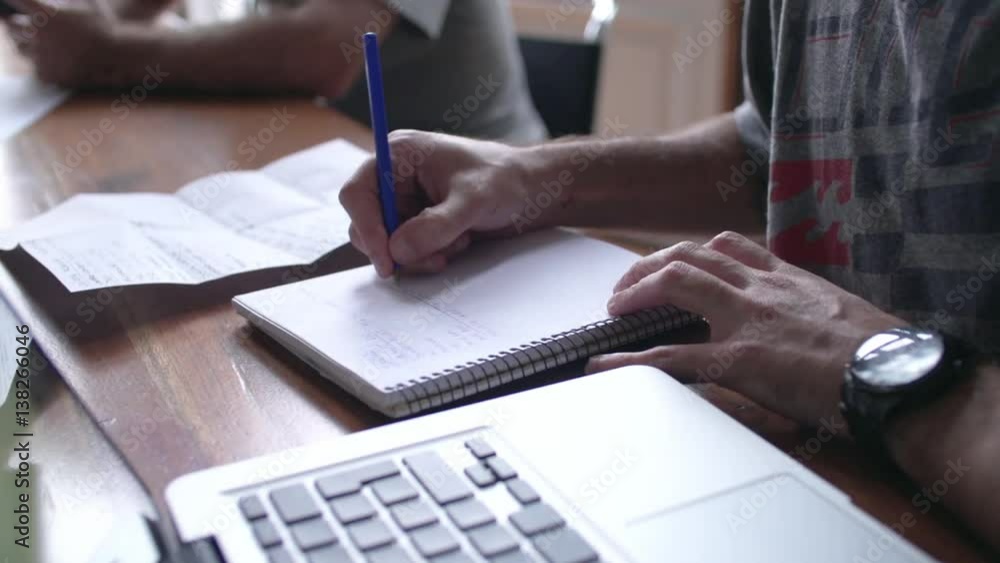 Close-up of man writing in notebook with laptop computer nearby