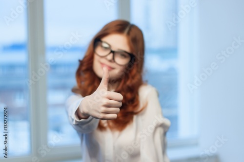 red-haired woman in glasses shows thumbs up on window background