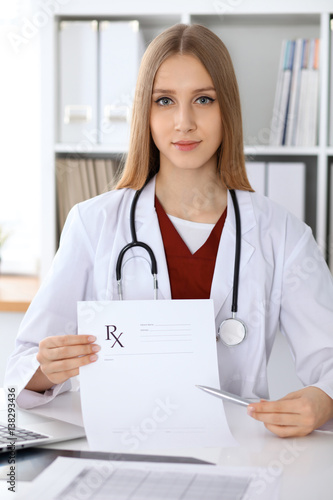  Young female doctor fills up medical form and pointing into medical prescription copy space area while looking at the camera. Health care and medicine concept