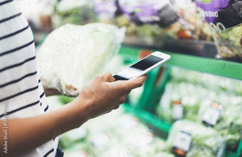 Young woman shopping healthy food in supermarket blur background. Female hands buy products cabagge using smartphone in store. Hipster at grocery using smartphone. Person comparing price of produce photo