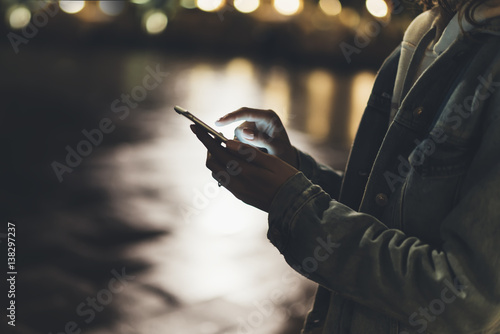 Girl pointing finger on screen smartphone on background illumination bokeh color light in night city, hipster using in female hands and texting mobile phone, mockup glitter street, content lifestyle