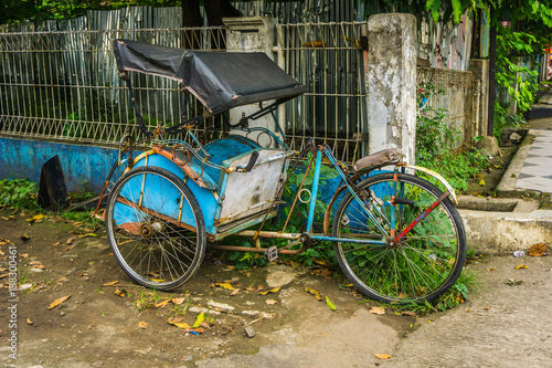 Blue pedicap or tricycle parked in side of the road near bush and wall with nobody around photo taken in Depok Indonesia photo