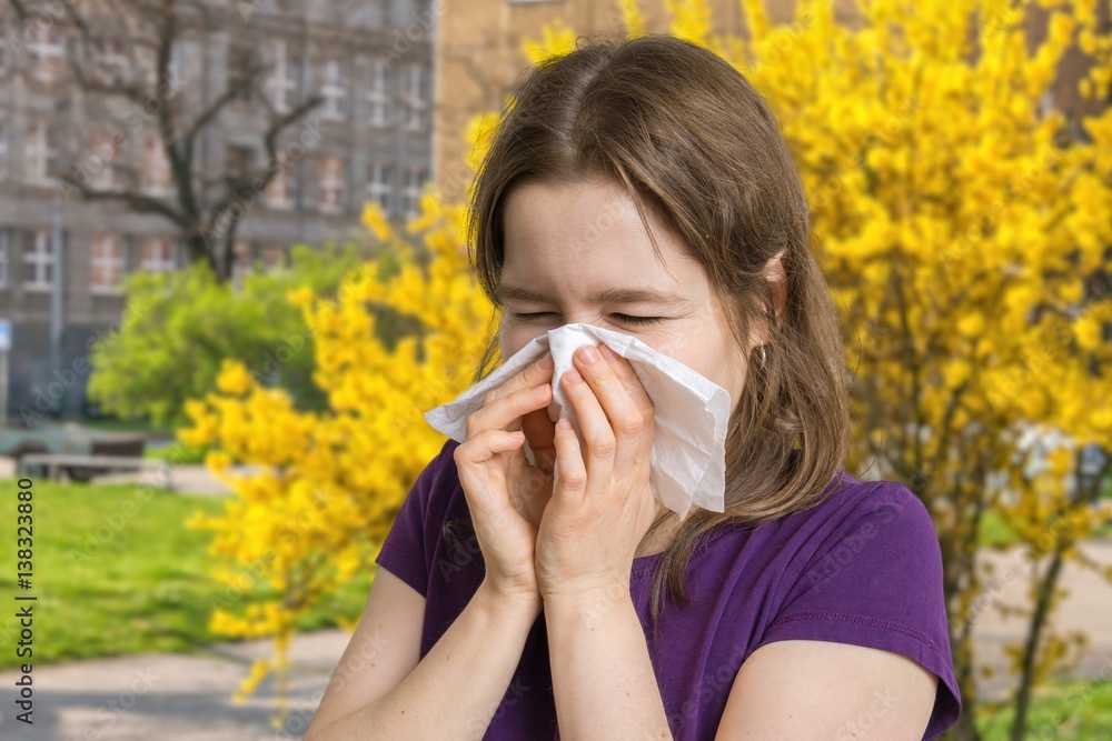 Allergy concept. Young allergic woman is sneezing and blowing her nose. Flowering trees in background. Allergy concept. Young allergic woman is sneezing and blowing her nose. Flowering trees in background.