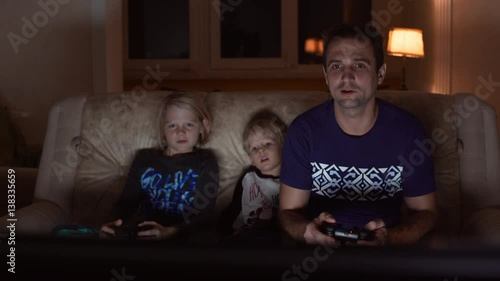 Handsome Caucasian father in blue t-shirt sitting at sofa with his two blonde sons playing console photo