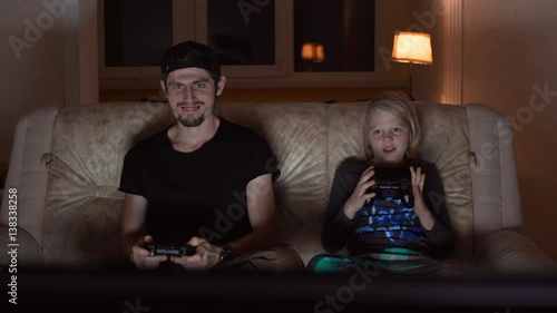 Adult Caucasian male with cap light bristle and black t-shirt sitting at sofa playing console with blonde young boy photo