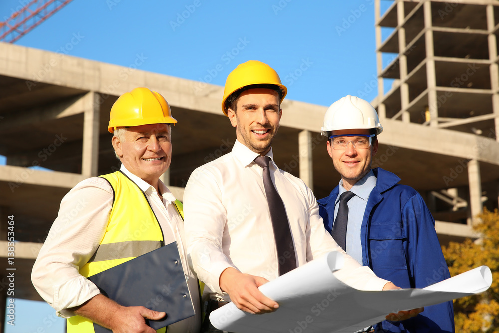 Engineer with drawing and workers standing against unfinished building Engineer with drawing and workers standing against unfinished building