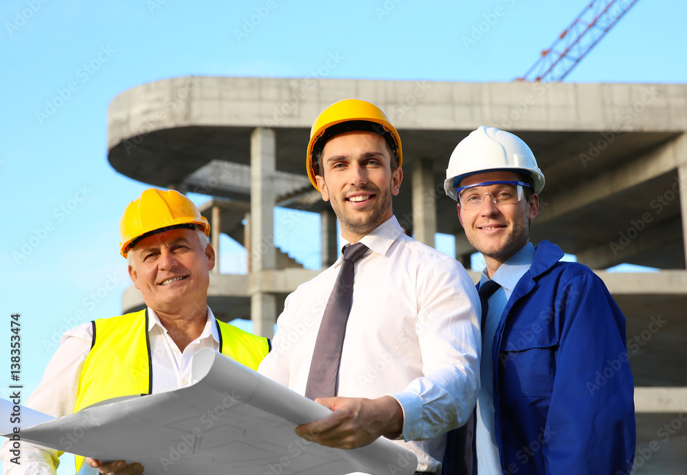 Engineer with drawing and workers standing against unfinished building Engineer with drawing and workers standing against unfinished building