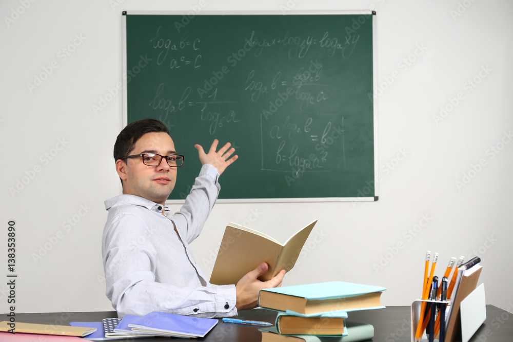 Handsome young teacher sitting at table in classroom Handsome young teacher sitting at table in classroom