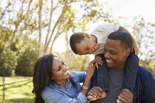 Parents Carrying Son On Shoulders As They Walk In Park