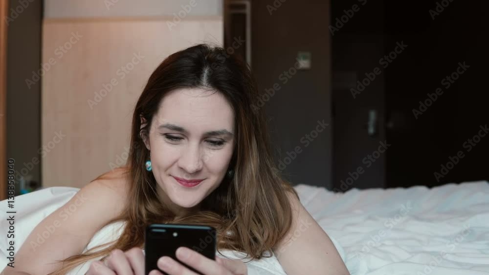Young woman using her smartphone while lying on the bed and smiling. Girl touches the screen and browsing the Internet. Young woman using her smartphone while lying on the bed and smiling. Girl touches the screen and browsing the Internet.