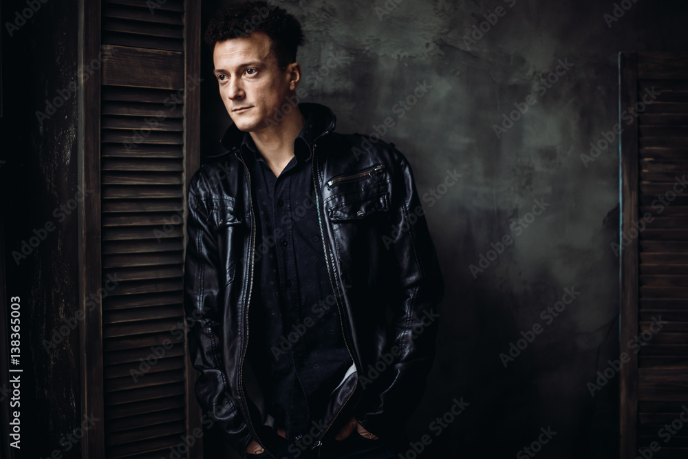 Handsome curly man in black jacket leans to wooden storm shutter Handsome curly man in black jacket leans to wooden storm shutter