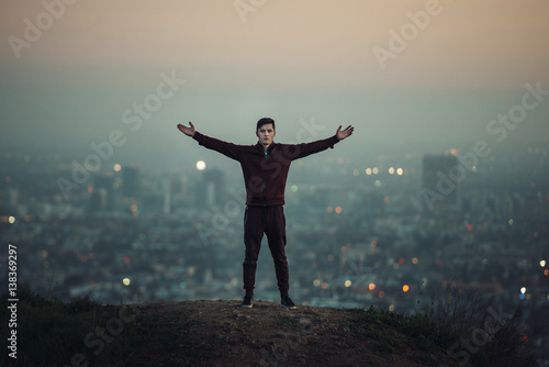 Tourist man standing on the hill with hands raised up next to big city light.