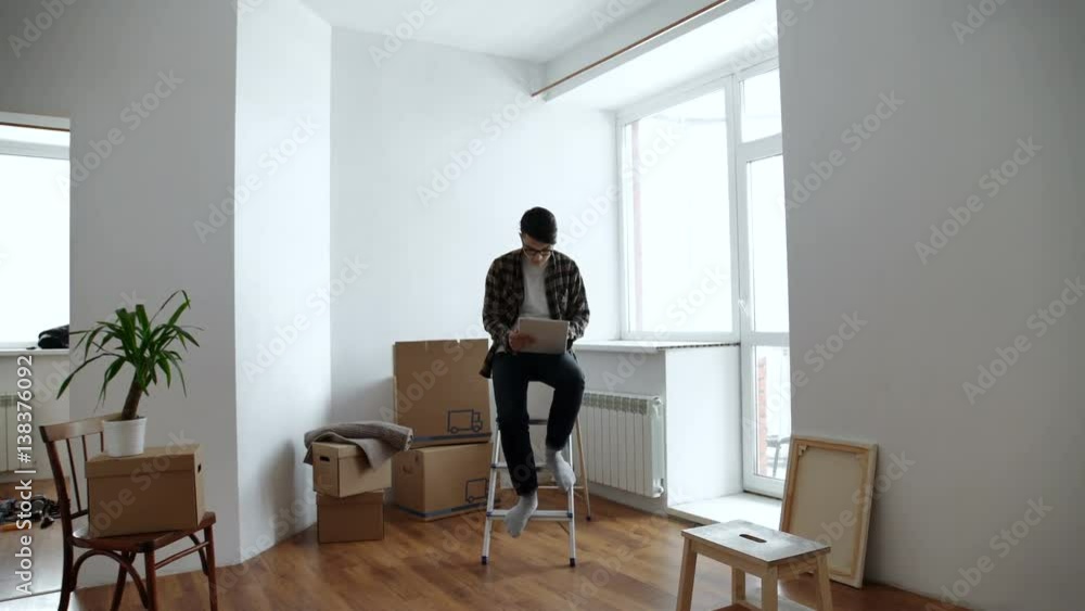 Smiling young man looking for some decoration ideas on a tablet. Moving into a new apartment. Cardboard boxes in the background. Smiling young man looking for some decoration ideas on a tablet. Moving into a new apartment. Cardboard boxes in the background.