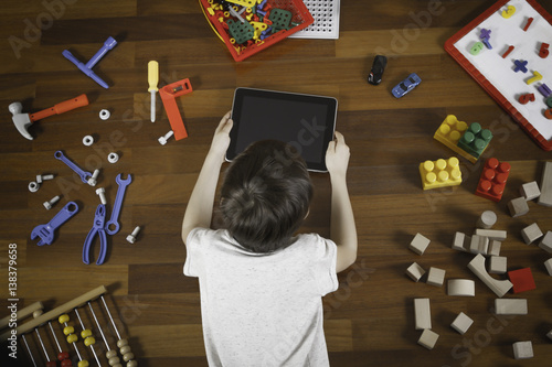 Little boy lying and playing with tablet computer. Lot of toys around him on the wooden floor. Top view