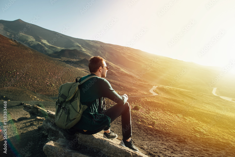 Man with rucksack sitting on rock at sunset on background of volcano Etna mountain in Sicily