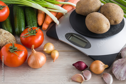 Potatoes on the weighing scales on the table with onions, tomatoes, cucumbers, garlic, carrots and other vegetables in the kitchen. Healthy eating and lifestyle.