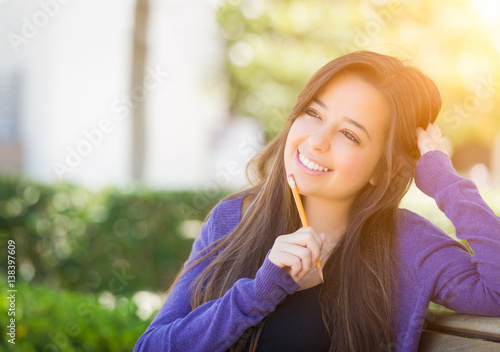 Attractive Pensive Mixed Race Female Student with Pencil Sitting on Campus Bench.
