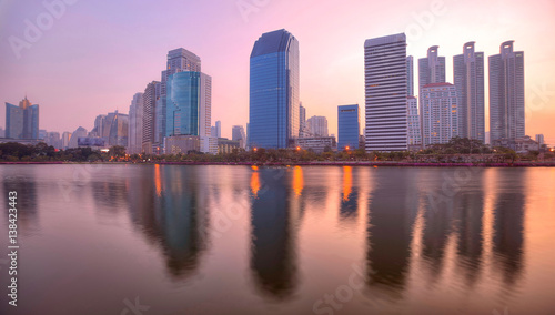 Beautiful city skyline of Bangkok at rosy dawn with lakeside skyscrapers and reflections ~ Morning view of modern buildings reflecting on smooth lake water in Benjakiti Park, Bangkok Thailand
