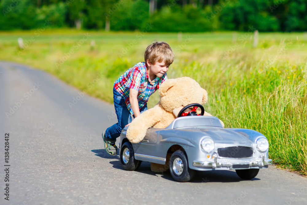 Little kid boy driving big toy car with a bear, outdoors.