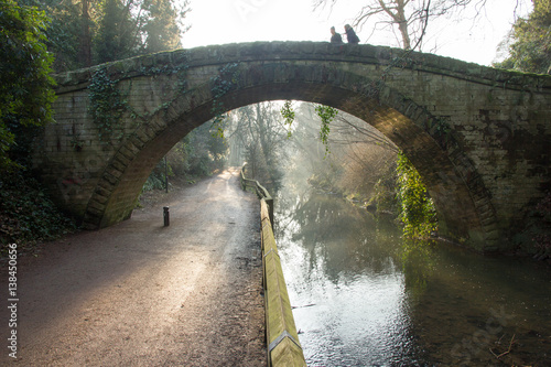 A couple walk across a solid stone hump back Victorian bridge as the golden wintry morning sun rays break through the trees and highlight the mist rising from the river