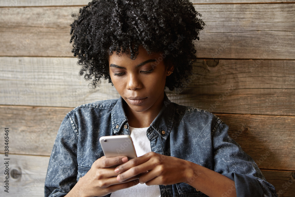 Beautiful young dark-skinned woman wearing trendy jeans jacket typing sms  on her generic smart phone, browsing newsfeed via social networks online  while waiting for friends at cafe with wooden walls Stock Photo
