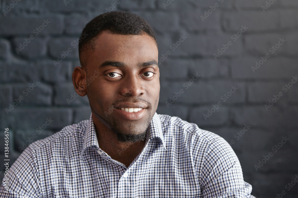 Close-up portrait of handsome young dark-skinned student or employee with small beard wearing formal checkered shirt looking and smiling at camera, sitting isolated against brick wall background Close-up portrait of handsome young dark-skinned student or employee with small beard wearing formal checkered shirt looking and smiling at camera, sitting isolated against brick wall background