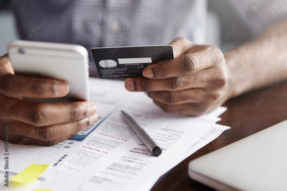Online payment and shopping concept. Cropped shot of African-American male holding cell phone in one hand and credit card in other, making transaction, suing mobile banking app during lunch at cafe Online payment and shopping concept. Cropped shot of African-American male holding cell phone in one hand and credit card in other, making transaction, suing mobile banking app during lunch at cafe