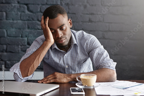 Frustrated tired young African-American employee touching his head, feeling absolutely exhausted because of overwork, calculating accounts, drinking another cup of coffee. Deadline and overwork photo