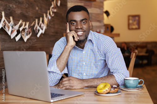 People, modern technology and communication concept. Cheerful dark-skinned male in formal shirt having rest at cafe during lunch break, sitting at table with mug, dessert and generic laptop pc