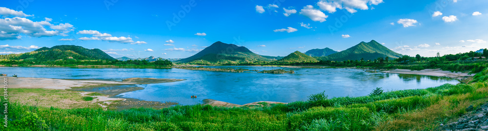 Panorama view of mountain full of green tree with river in front and clear blue sky.