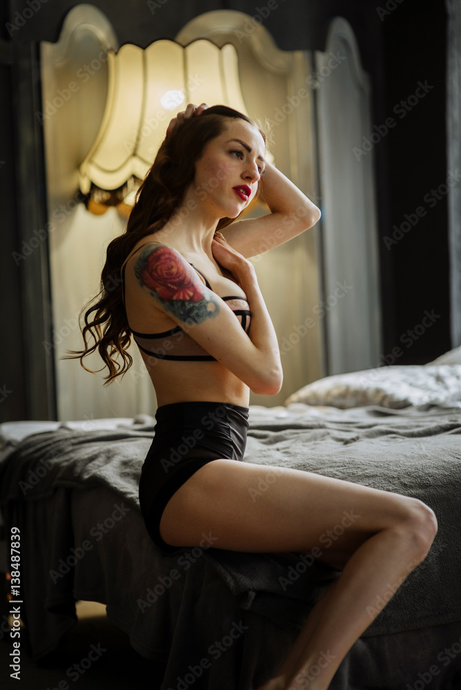 Fototapeta premium Boudoir photo of sexy girl with tattoo and long hair wearing stylish black lingerie posing in the bedroom. Woman underwear present for man.