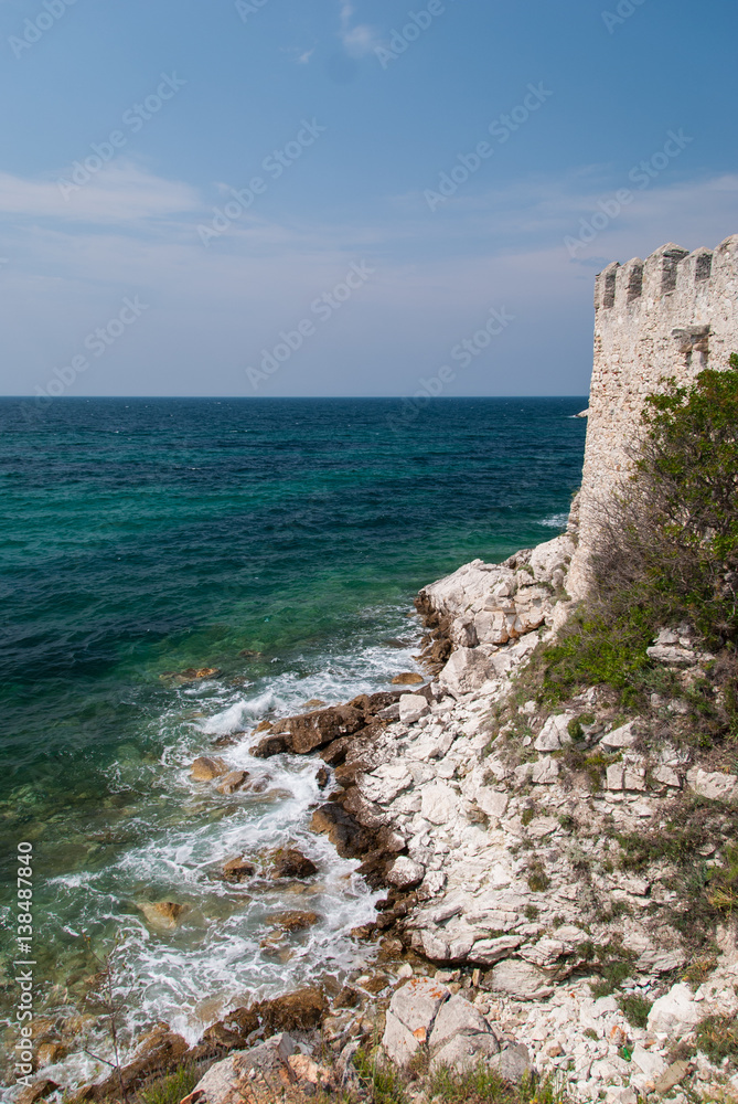 Ancient stone fortress wall on the edge of the Mediterranean sea with waves hitting large stones at the bottom of the cliff