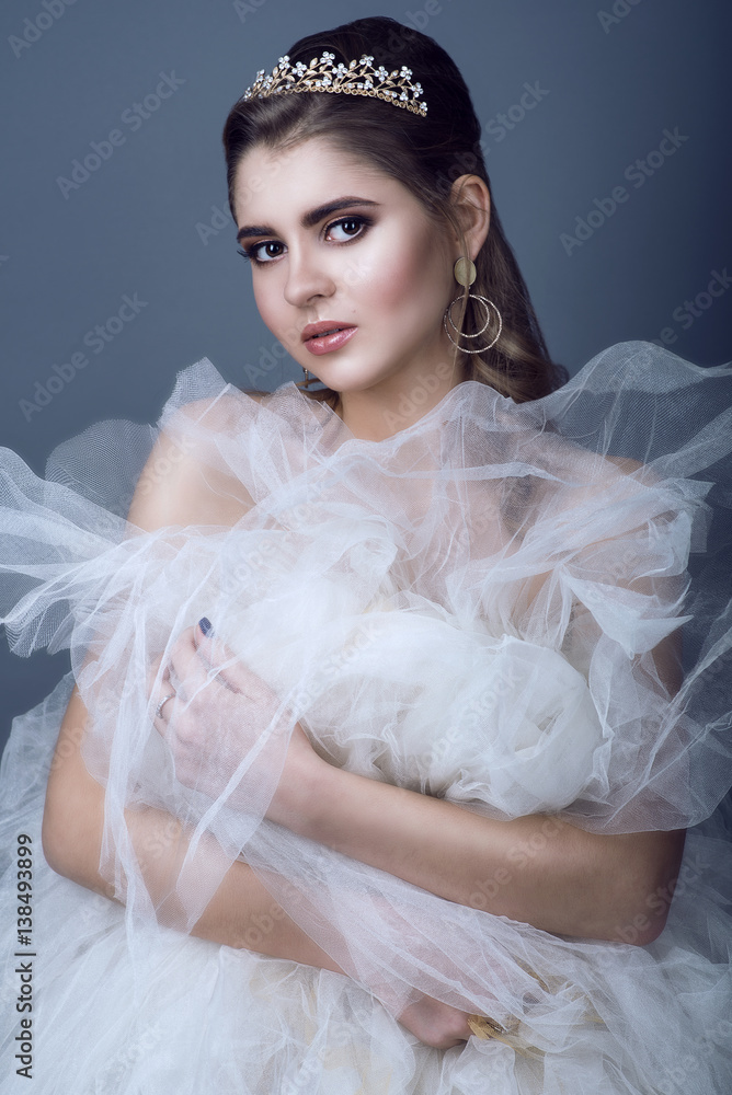 Portrait of young beautiful bride in diadem and earrings with naked shoulders pressing fluffy skirt of her wedding dress to her breast with wistful gaze. Isolated on grey background. Studio shot
