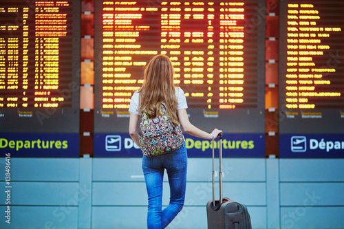 Tourist girl with backpack and carry on luggage in international airport, near flight information board