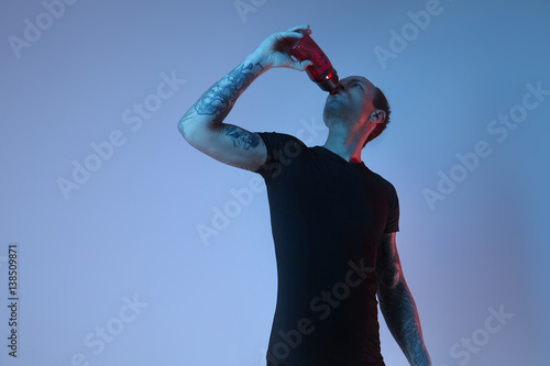 Fashion portrait of sport fit attractive man drinking water from bottle, tattooed hands, hipster look. Color flash studio light