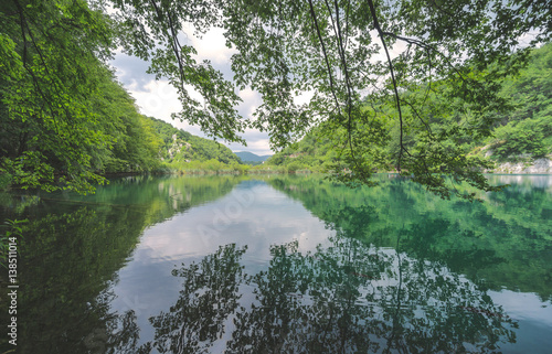 Fototapeta Naklejka Na Ścianę i Meble -  Trees, sky and mountains are reflected in the crystal clear lakes of Plitvice Lakes National Park, with unrecognizable tourists walking along the boardwalk in the distance.