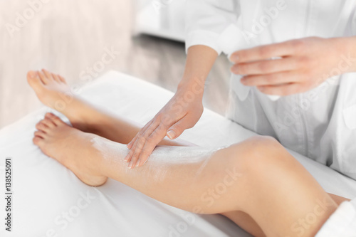 Beautician applying moisturizing cream on female legs in spa center