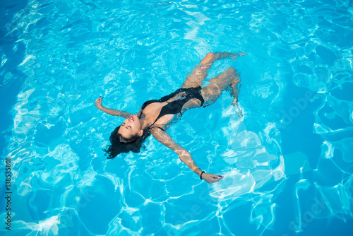 Attractive woman in a black bathing suit floating on her back in the swimming pool and relaxing, with copy space