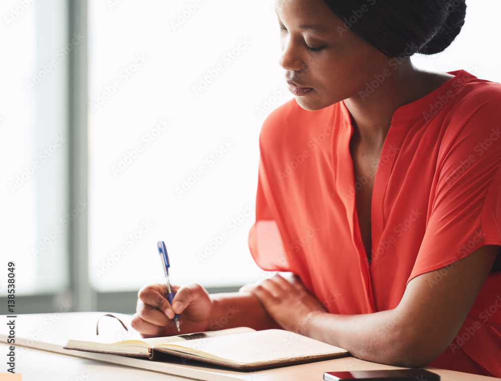 Beautiful young african american writer busy making hand written notes while wearing a bright orange blouse and sitting next to large bright windows. Beautiful young african american writer busy making hand written notes while wearing a bright orange blouse and sitting next to large bright windows.