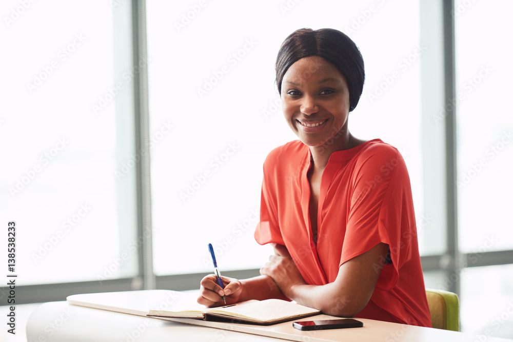 Female African American writer wearing an orange blouse while looking into the camera still holding a pen in her hands that she was using to write down her notes. Female African American writer wearing an orange blouse while looking into the camera still holding a pen in her hands that she was using to write down her notes.
